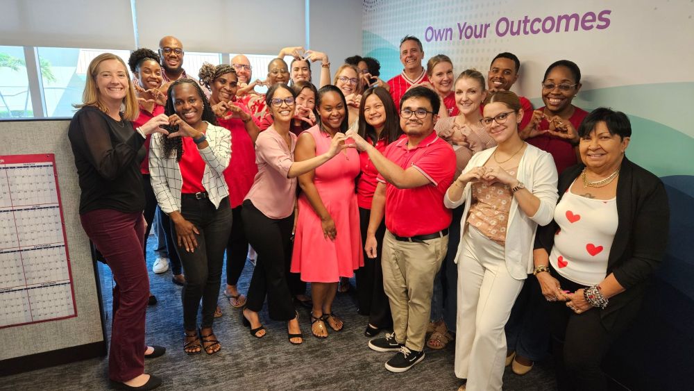 The GCM team poses with their hands in heart shapes in front of a wall that says Own Your Outcomes